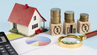 Model house next to stacked coins on wooden blocks, financial charts, and magnifying glass symbolising home savings and investment planning