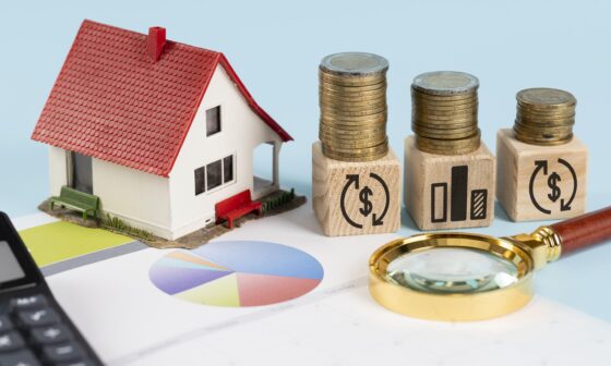 Model house next to stacked coins on wooden blocks, financial charts, and magnifying glass symbolising home savings and investment planning