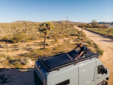 Person installing a 200W flexible solar panel on the roof of a camper van in a sunny desert landscape.